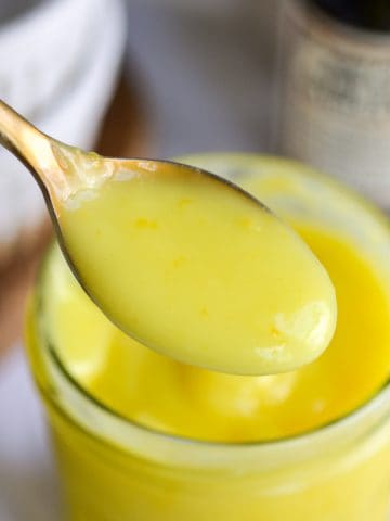 A spoon with vegan lemon curd on it over a glass jar filled with lemon curd.