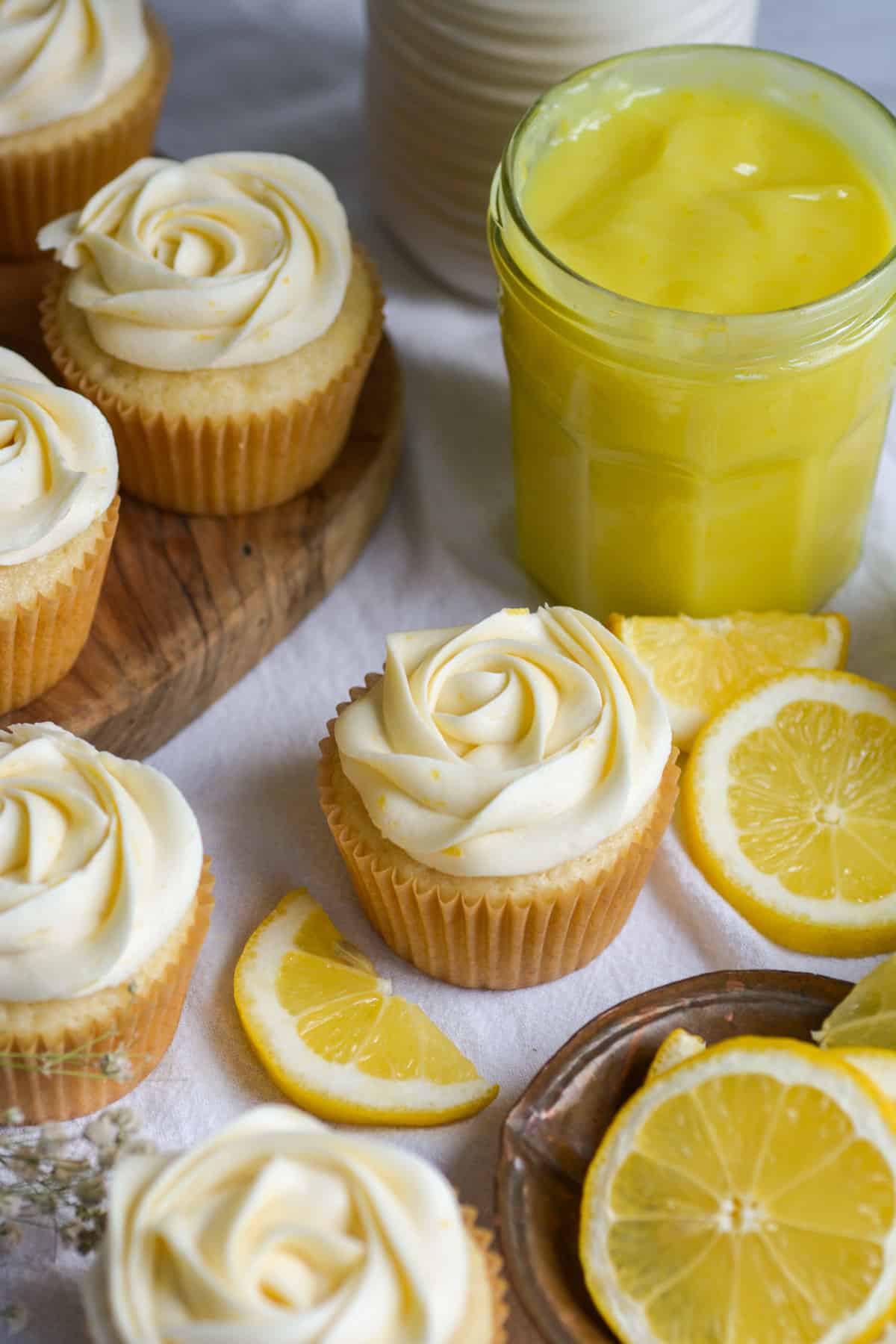 Frosted cupcakes on a cloth with lemon slices in the foreground and a glass jar of lemon curd in the background.