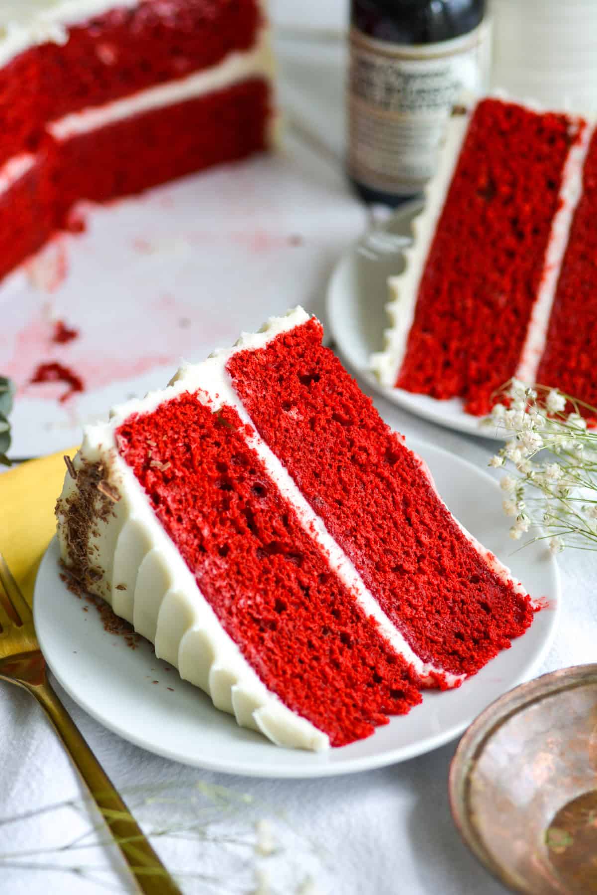 Vegan red velvet cake slice on a small plate with another cake slice behind it. the remaining cake is in the background.