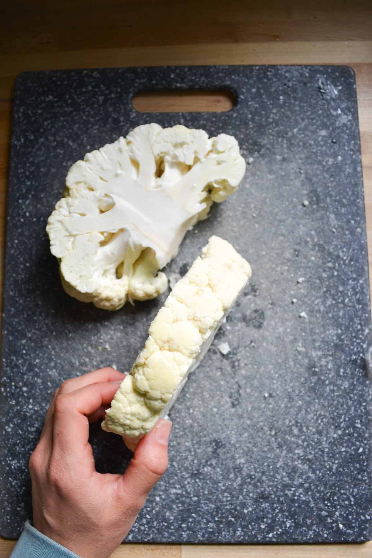 A hand holding a vegan cauliflower steak to show the thickness with another cauliflower steak in the background on a cutting board.