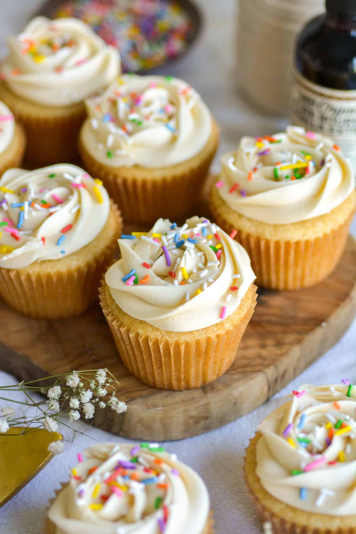 Vegan Vanilla Cupcakes topped with rainbow sprinkles on a small wooden board with cupcakes in the foreground.
