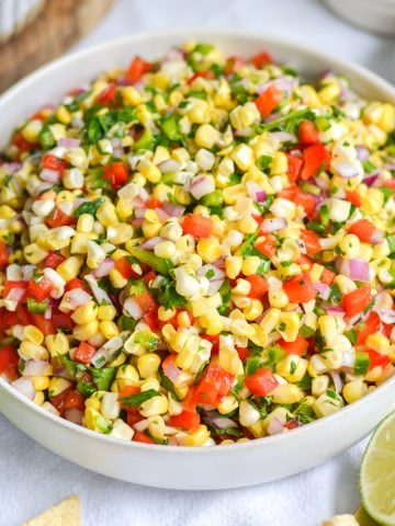 A wide bowl full of Easy corn salsa on a white cloth with a lime and a jalapeño in the foreground.