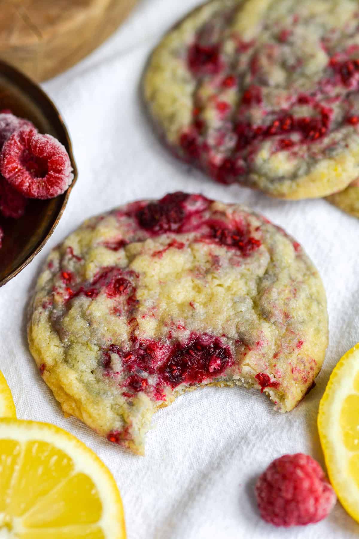 Vegan Lemon Raspberry Sugar Cookie with a bite taken out of it on a white cloth with lemon slices and frozen raspberries in the scene.