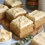Squares of vegan banana cake topped with peanut butter frosting on a small wooden board with a bowl of frosting in the foreground.