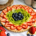 Vegan Fruit Tart topped with strawberries, blueberries and kiwi slices on a white cloth with strawberries in the foreground.