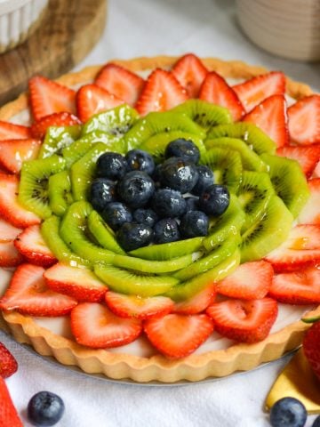 Vegan Fruit Tart topped with strawberries, blueberries and kiwi slices on a white cloth with strawberries in the foreground.