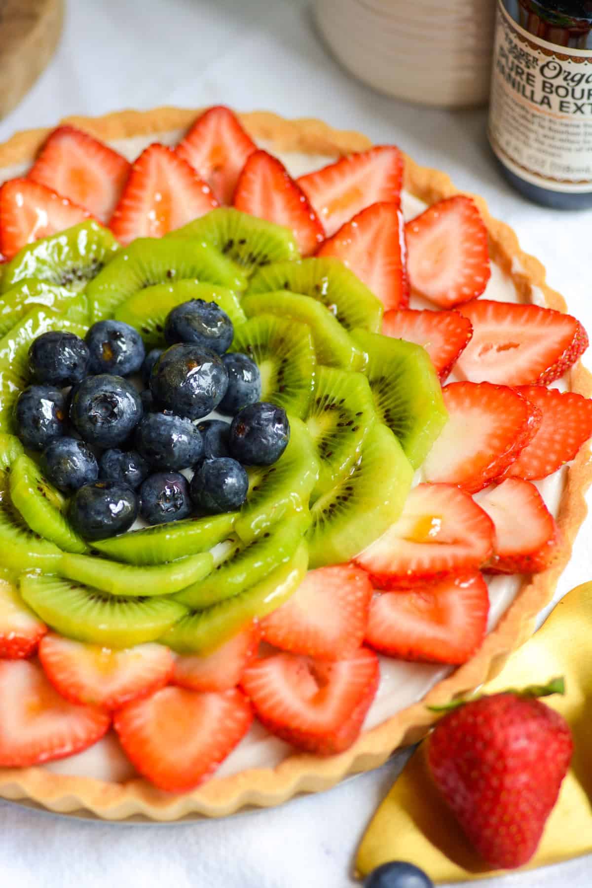 Vegan fruit tart that is topped with sliced strawberries, kiwi and blueberries on a white cloth with a strawberry in the foreground.