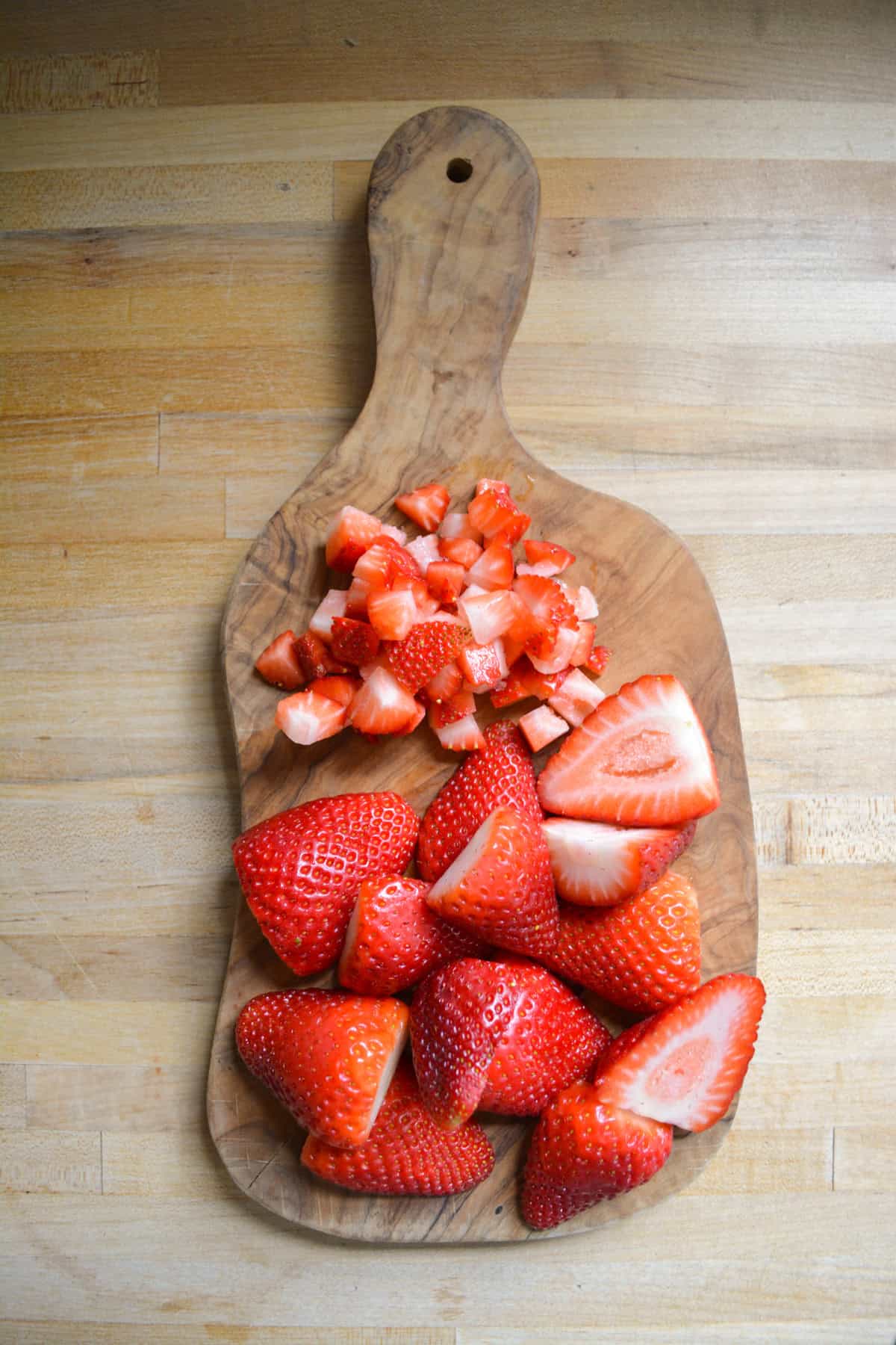 Strawberries on a small wooden cutting board. Some are sliced in half and some are chopped.