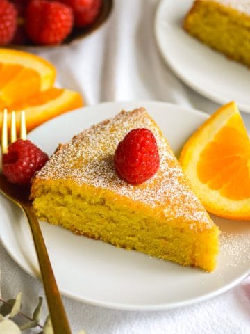 A slice of Vegan Orange Olive Oil Cake on a small plate with a raspberry on top and and orange slice on the side.