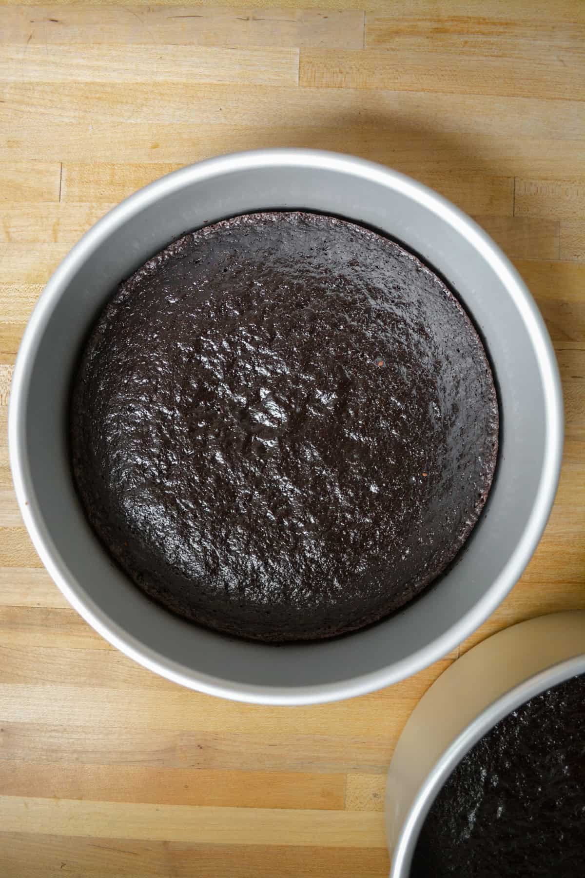 Baked chocolate cakes in their cake pans on a wooden surface.