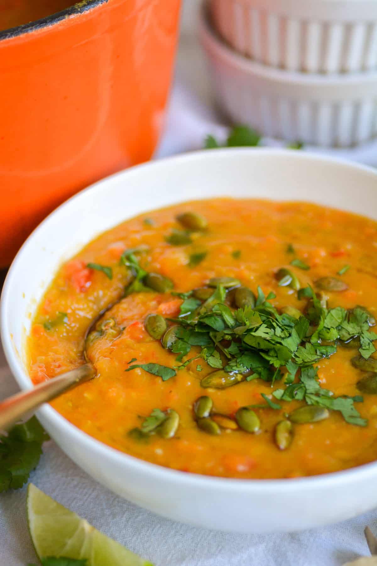 Carrot Red Lentil Soup topped with chopped cilantro and toasted pumpkin seeds in a bowl with a spoon in it.