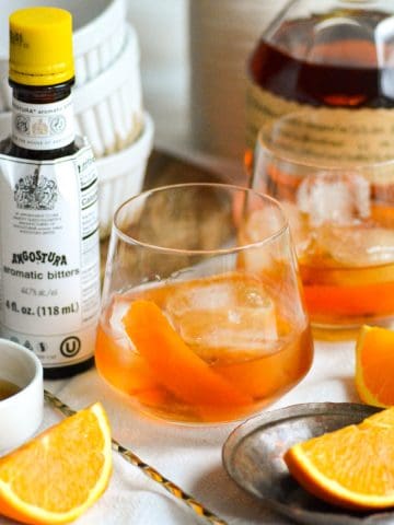 A Maple Old Fashioned in a glass with orange slices in the scene and a bottle of Blanton's bourbon in the background.