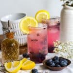 Blueberry Lavender Mocktail in a tall glass garnished with a lemon wheel which a sliced lemon and bowl of blueberries on the table.
