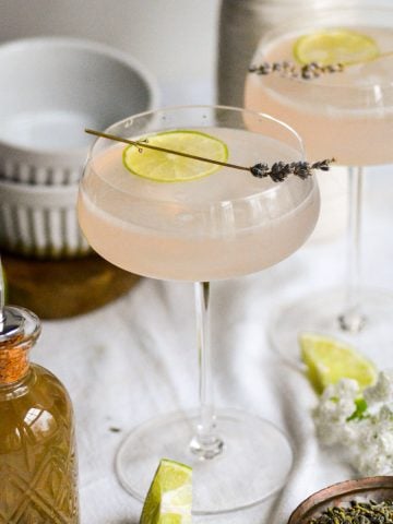 Two Lavender Gimlets in glasses on a linen cloth with a bottle of lavender syrup in the scene.