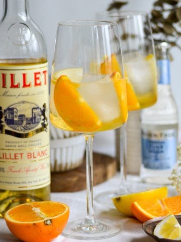 Lillet Blanc Spritz in a wine glass with ice cubes and orange slices with a bottle of Lillet in the background.