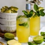 Two Pineapple Mojito Mocktails garnished with mint with limes and sliced pineapple on the table.