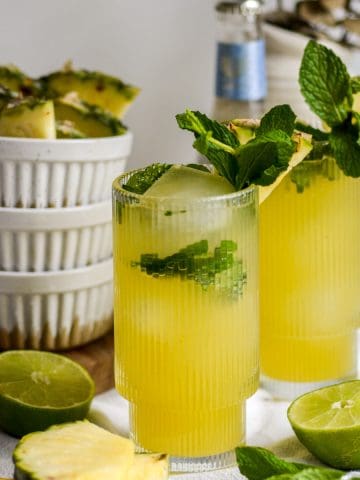 Two Pineapple Mojito Mocktails garnished with mint with limes and sliced pineapple on the table.