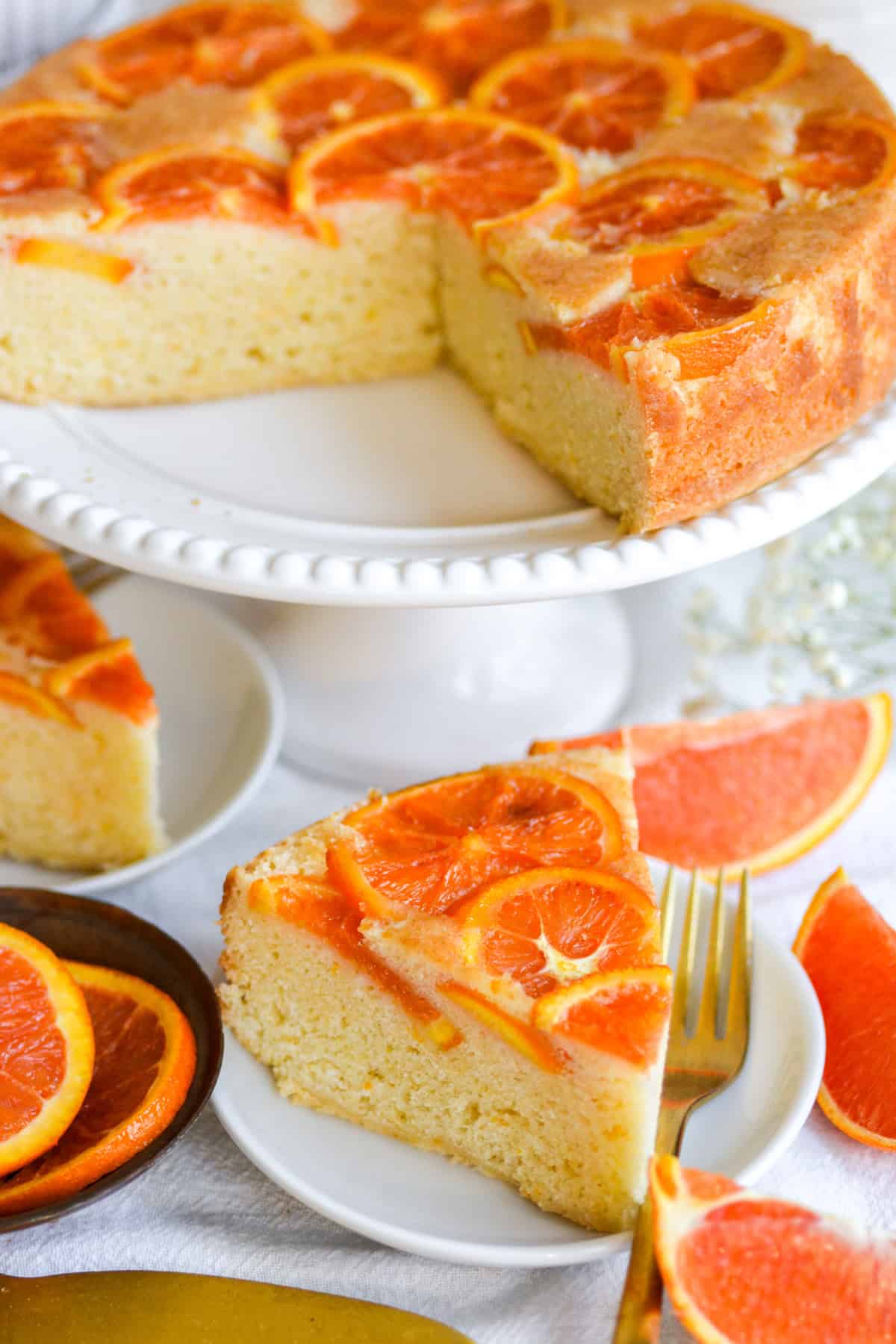 A slice of Orange Upside Down Cake on a small plate with the rest of the cake on a cake stand in the background.