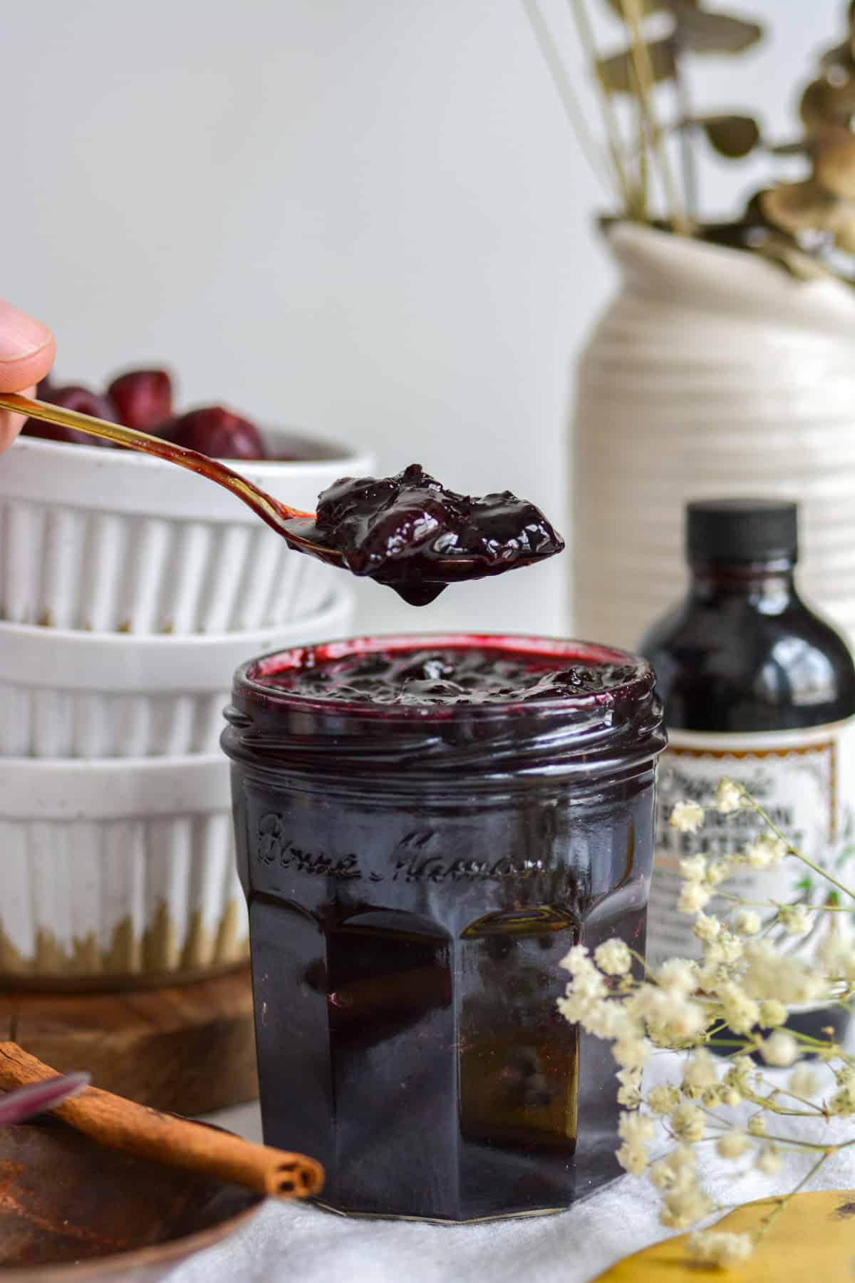 A spoon with a scoop of cherry jam hovering over a glass jar of cherry jam.