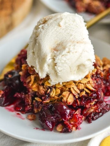 A scoop of cherry crisp with frozen cherries on a small plate topped with a scoop of ice cream.