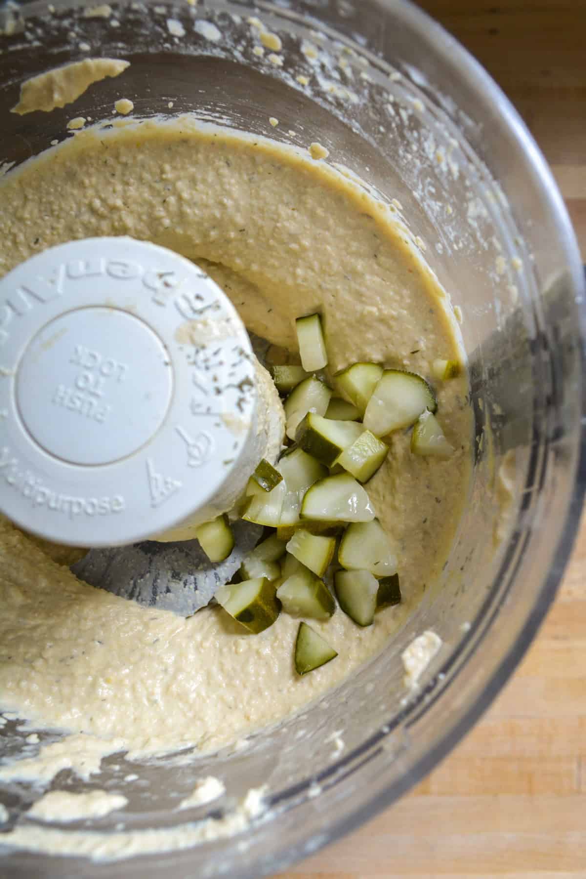 Chopped pickles added to hummus in the bowl of a food processor.