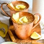 Two Apple Cider Mule Mocktails garnished with a cinnamon stick and apple slices on a wooden board.