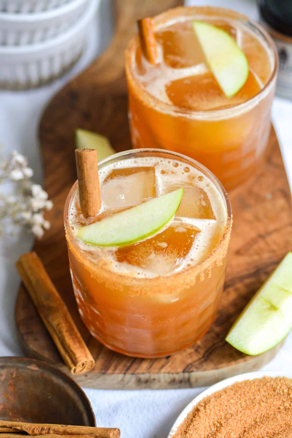 Two Bourbon Whiskey Apple Cider Cocktails garnished with cinnamon sticks on a wooden board.