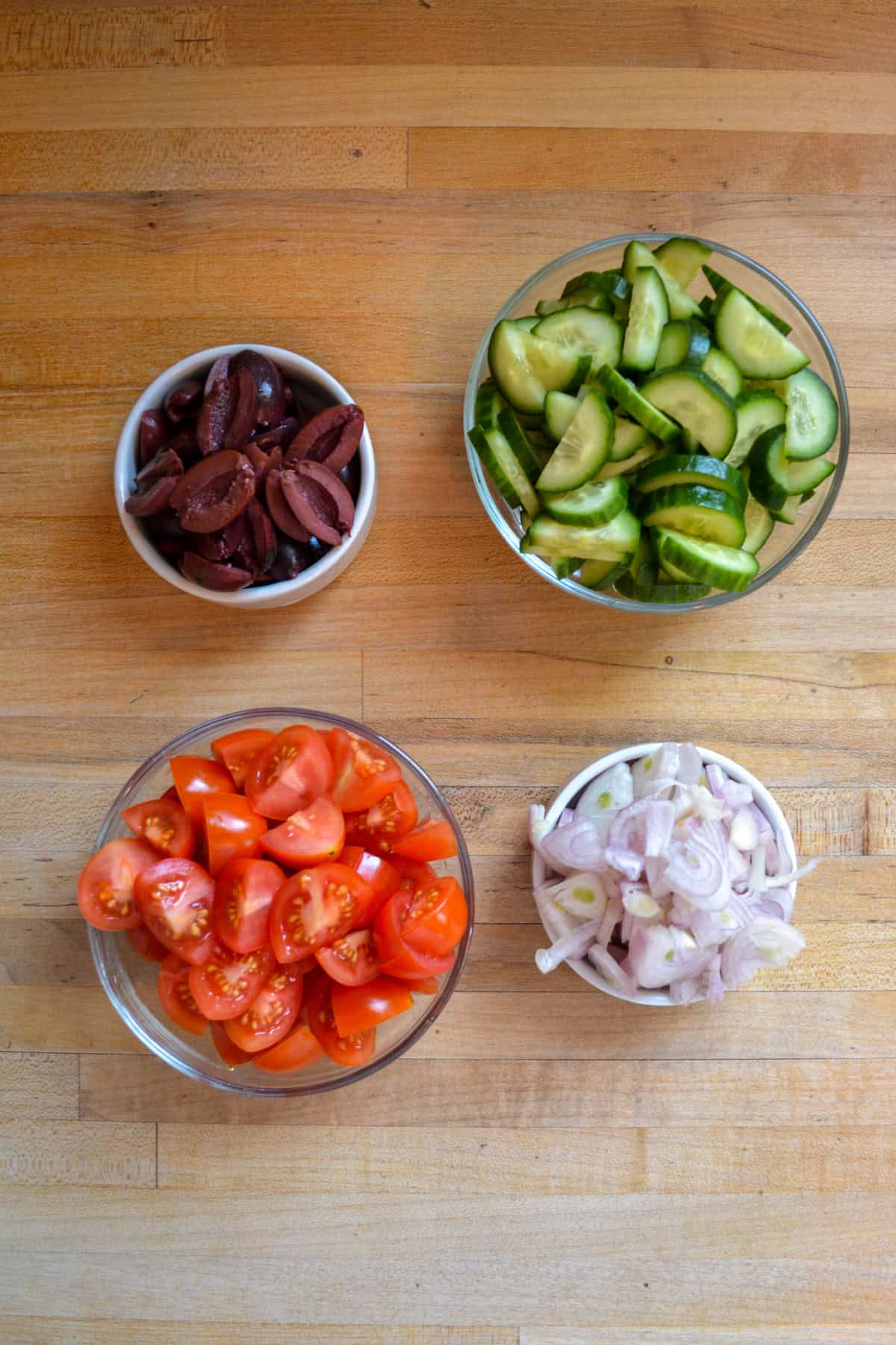 Chopped tomatoes, cucumber, shallot and olives in small bowls on a wooden surface.
