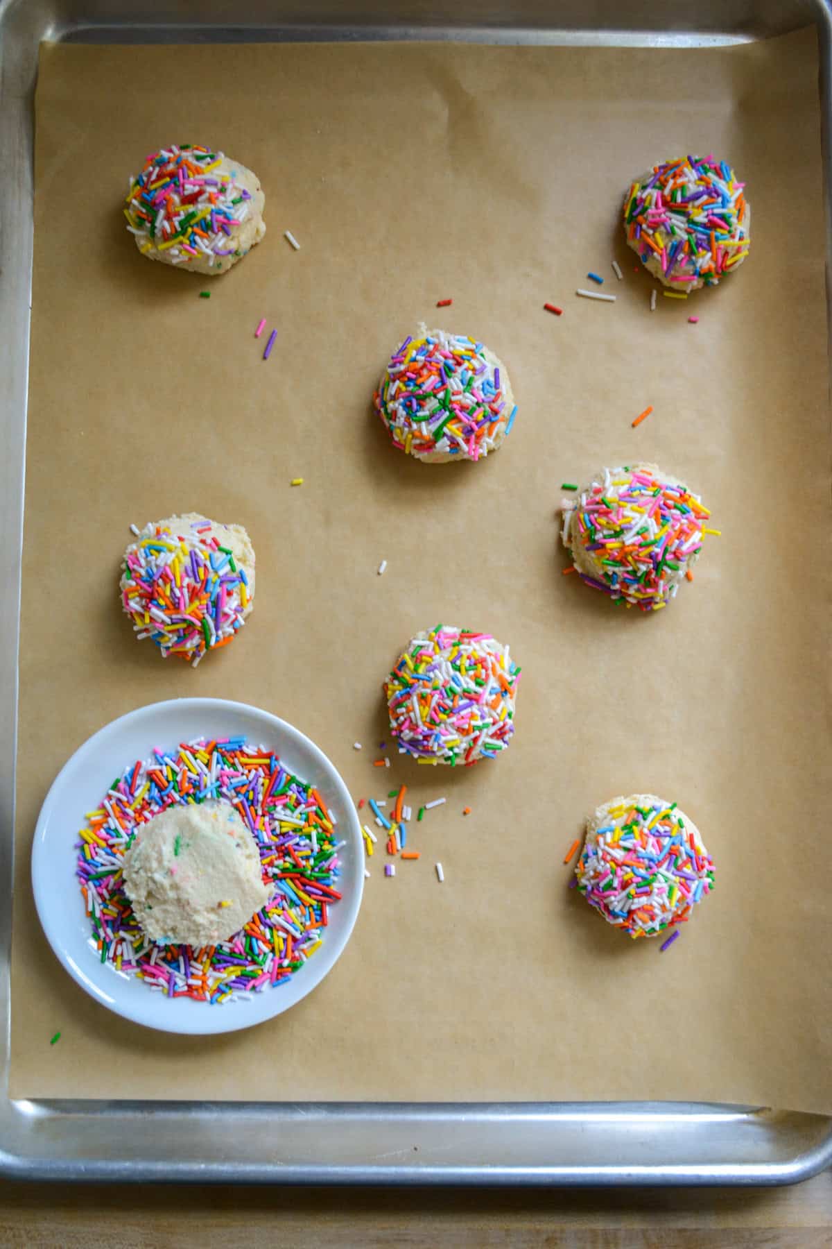 Dunking the cookie dough balls in sprinkles and placing them on a parchment-lined baking sheet.