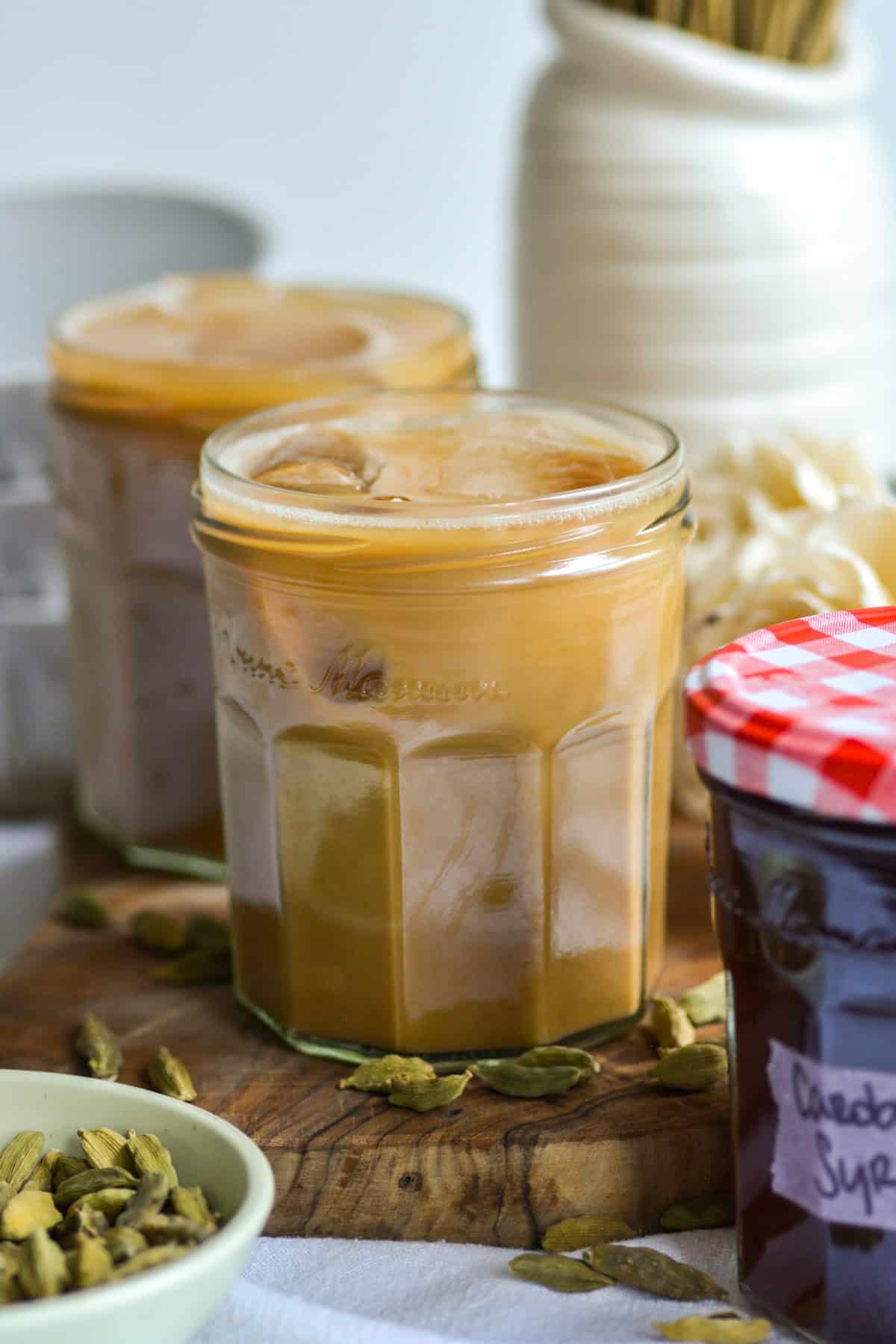 Two glasses of cardamom latte on a wooden board with cardamom pods in the foreground.