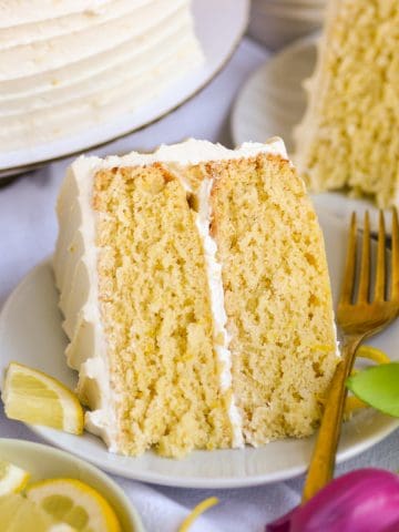 A slice of Vegan Lemon Cake on a plate next to a bowl of sliced lemons.