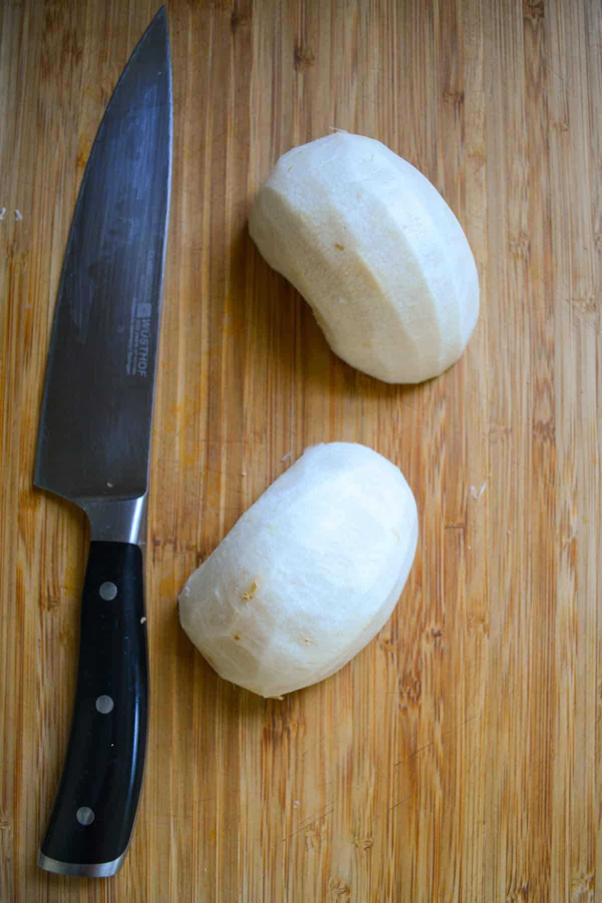 A peeled and halved jicama on a wooden cutting board next to a chef's knife.
