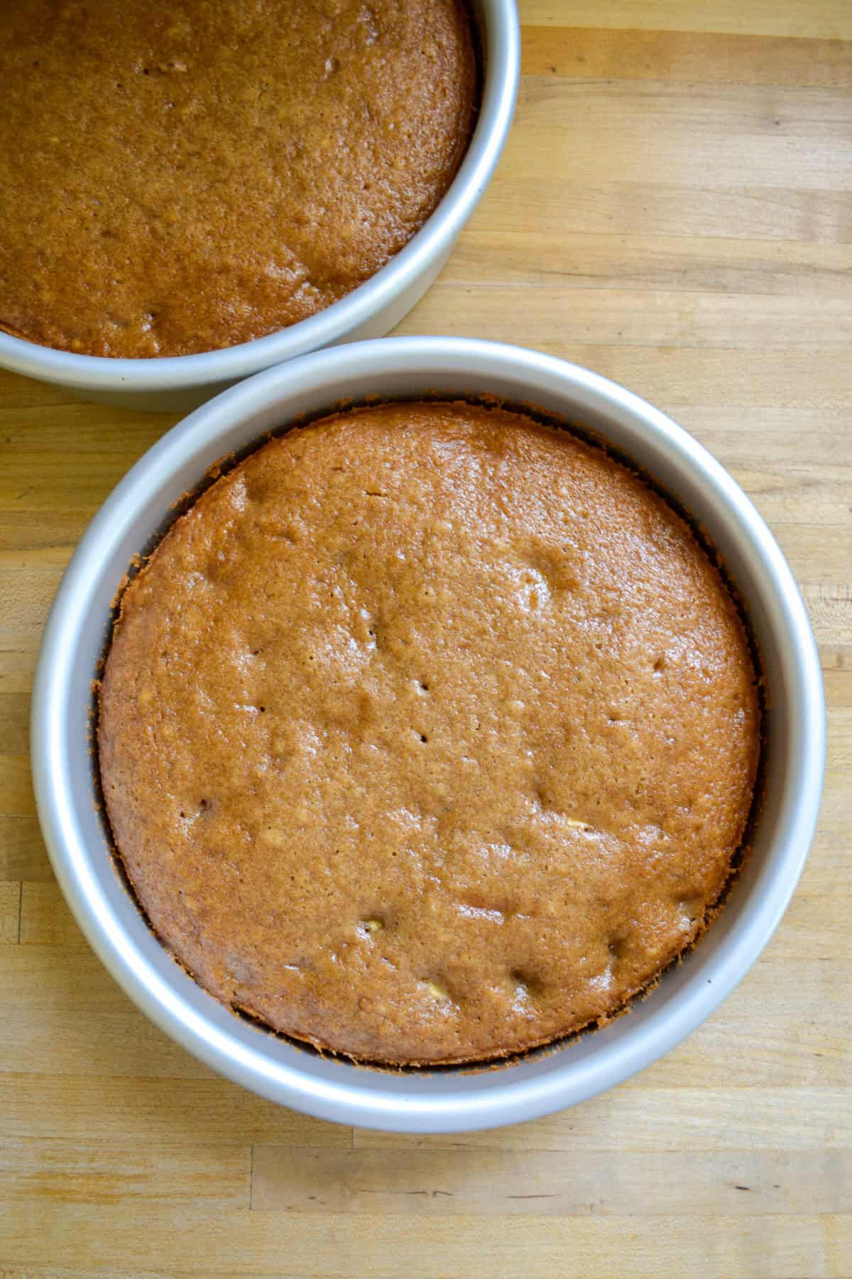 Baked cake layers in their cake pans on a wooden surface.